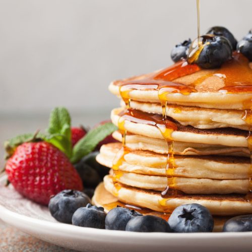 Close-up delicious pancakes, with fresh blueberries, strawberries and maple syrup on a light background. With copy space. Sweet maple syrup flows from a stack of pancake.